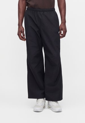 Homme portant un pantalon noir ample et des baskets blanches, debout devant un fond clair uni.