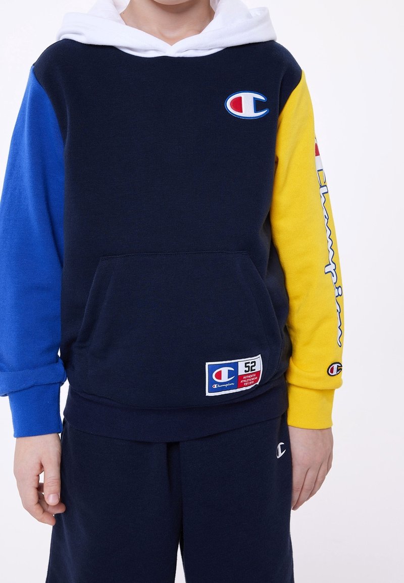 Sweat à capuche bicolore bleu marine et bleu, manches jaunes, poche avant et capuche blanche ; présente le logo Champion et une étiquette tissée.