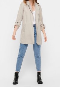 Kvinna i beige lång blazer över en vit topp, blå högmidjade jeans och svarta ankelstövlar, står med benen isär mot en vit bakgrund.