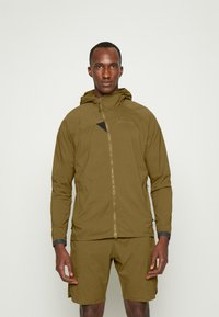 Khaki lightweight jacket with a zip front, hood, and triangular black accent; paired with matching shorts, showcasing a smooth texture.