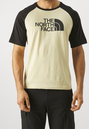 Mężczyzna w beżowo-czarnym T-shircie z raglanowymi rękawami z logo „The North Face” na klatce piersiowej i czarnych spodniach typu cargo.