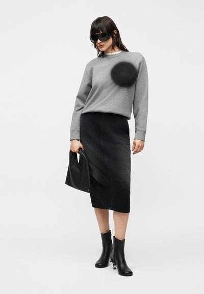 Γυναίκα που φοράει υπερμεγέθη μαύρα γυαλιά ηλίου, γκρι φούτερ, μαύρη φούστα μέχρι τα γόνατα, μαύρες μποτάκια μέχρι τον αστράγαλο, κρατώντας μαύρη τσάντα.