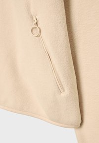 Beige fleece-stof med en glat tekstur, der har en sidelomme med lynlås og en ringtræk. Nederkanten er rundet med en subtil syning.