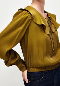 Blouse en satin vert olive avec grand col, manches longues bouffantes et ourlet élastiqué, portée avec un jean bleu foncé.