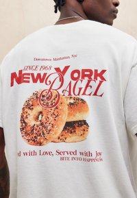 Balta kokvilnas T-kreklā ir grafiska apdruka ar bagelēm melnā un brūnā krāsā, kā arī sarkans teksts ar uzrakstiem "NEW YORK BAGEL" un "Since 1968."