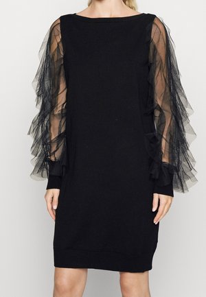 Schwarzes knielanges Kleid mit transparenten, geschichteten Rüschenärmeln und rundem Ausschnitt, getragen von einer stehenden Person.