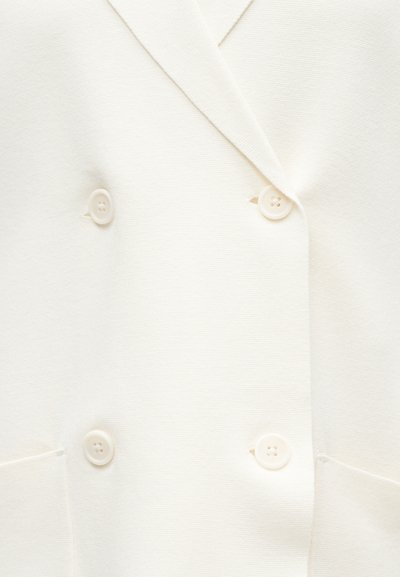 Κοντινό λευκού διπλού σακακιού με τέσσερα ταιριαστά κουμπιά και δύο εμπρόσθιες τσέπες πάνω σε υφασμάτινη υφή.