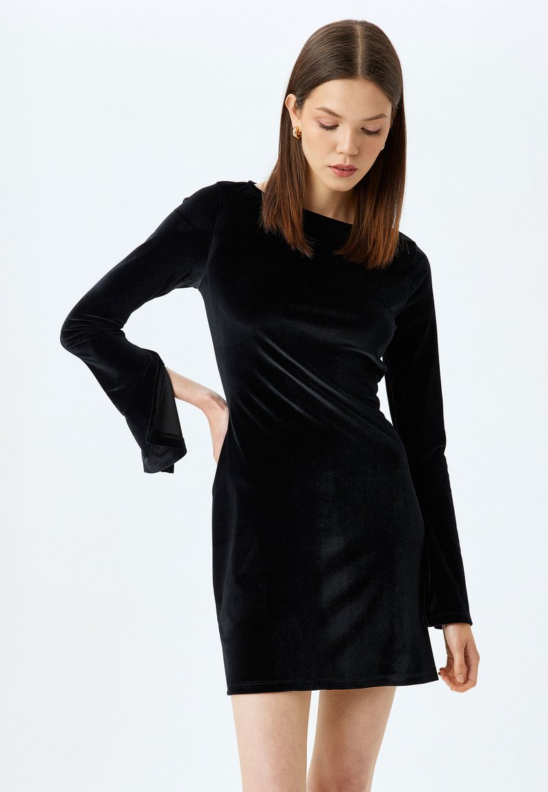 Vestido negro de terciopelo de manga larga con una silueta ajustada, que presenta puños acampanados y un escote redondo. Textura suave, con longitud a mitad del muslo.