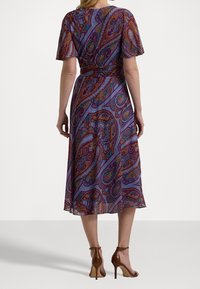 Robe paisley violette à manches courtes, longueur genou, tissu fluide, motifs détaillés en rouge et bleu, associée à des sandales à talons hauts marron.