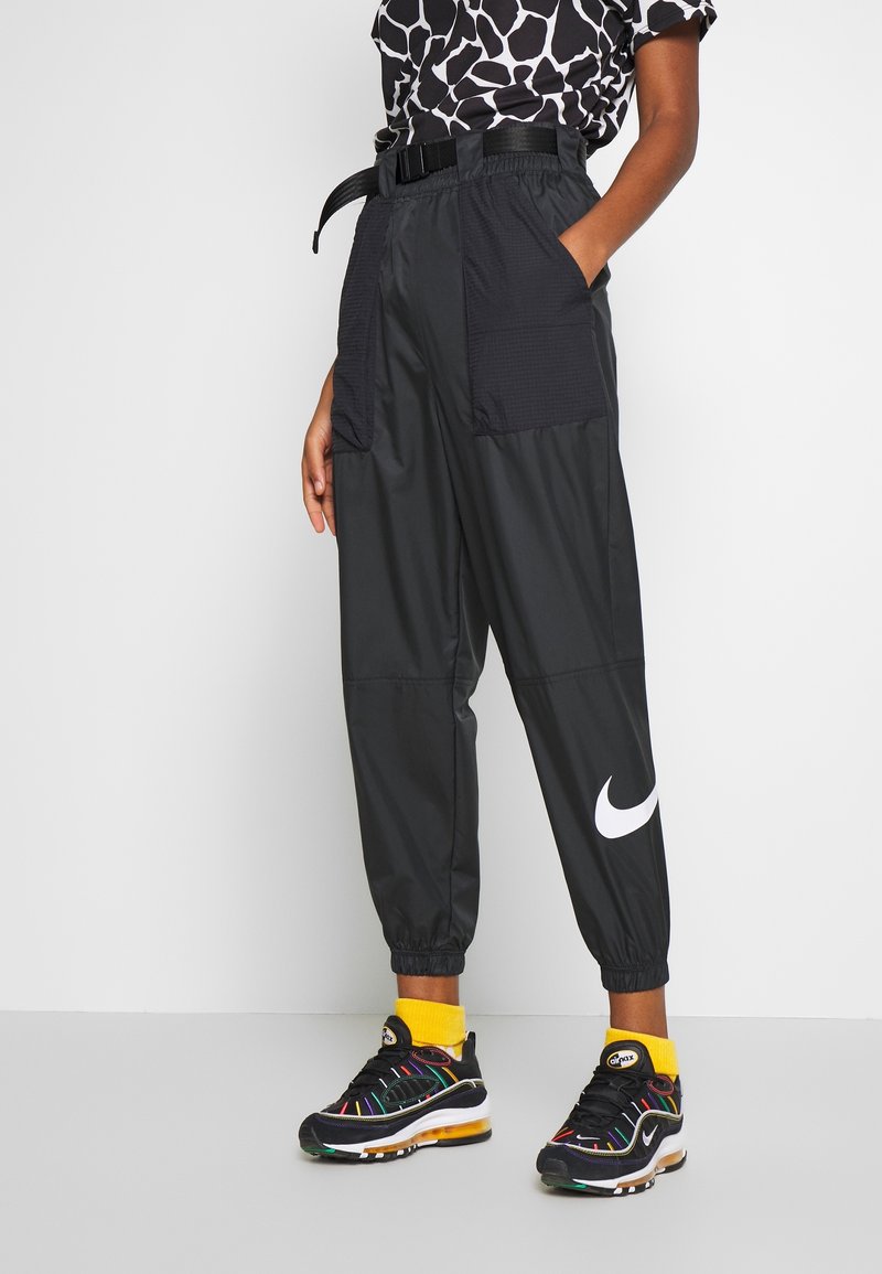 Svarta Nike-joggers med bälte i midjan, två stora fickor med texturerade paneler och elastiska manschetter. Bärs med färgglada Nike-sneakers.