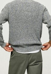 Grauer Strickpullover mit strukturiertem Muster, mit gerippten Bündchen und Saum. Getragen über einem weißen Hemd, kombiniert mit olivgrünen Hosen.