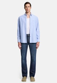 Ljusblå skjorta med lång ärm och knappar, vit linne under, parat med mörkblå jeans och beige skor. Klassisk passform, minimal detaljer.