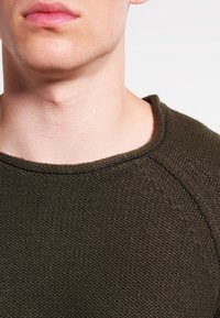 Olive zöld, textúrált pulóver, kerek nyakkivágással és finom varrási részletekkel, puha kötött anyagból készült.