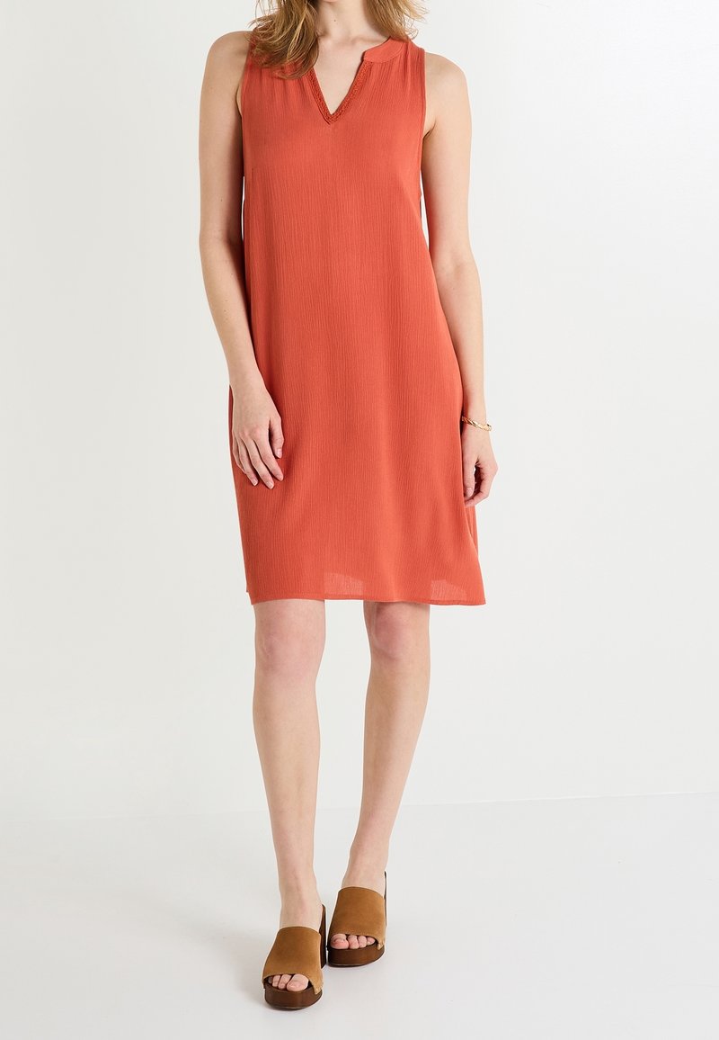 Femme portant une robe orange sans manches longueur genoux avec un col en V et des sandales compensées ouvertes à l'avant marron, se tenant devant un fond blanc.