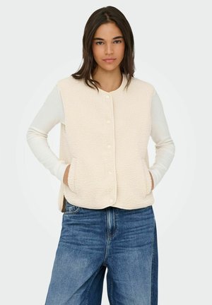 Beige fleece vest with a soft texture, round neckline, button closure, and side pockets, layered over a white long-sleeve top and blue jeans.