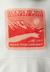 Logo brodé rouge et pêche sur fond blanc, représentant des montagnes, un soleil et le texte "NAPAPIJRI FAIS TA PROPRE ROUTE ©1987."