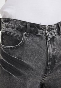 Gros plan sur un jean en denim noir délavé porté avec une chemise blanche côtelée rentrée, montrant les détails de la poche avant, des passants de ceinture et du bouton.