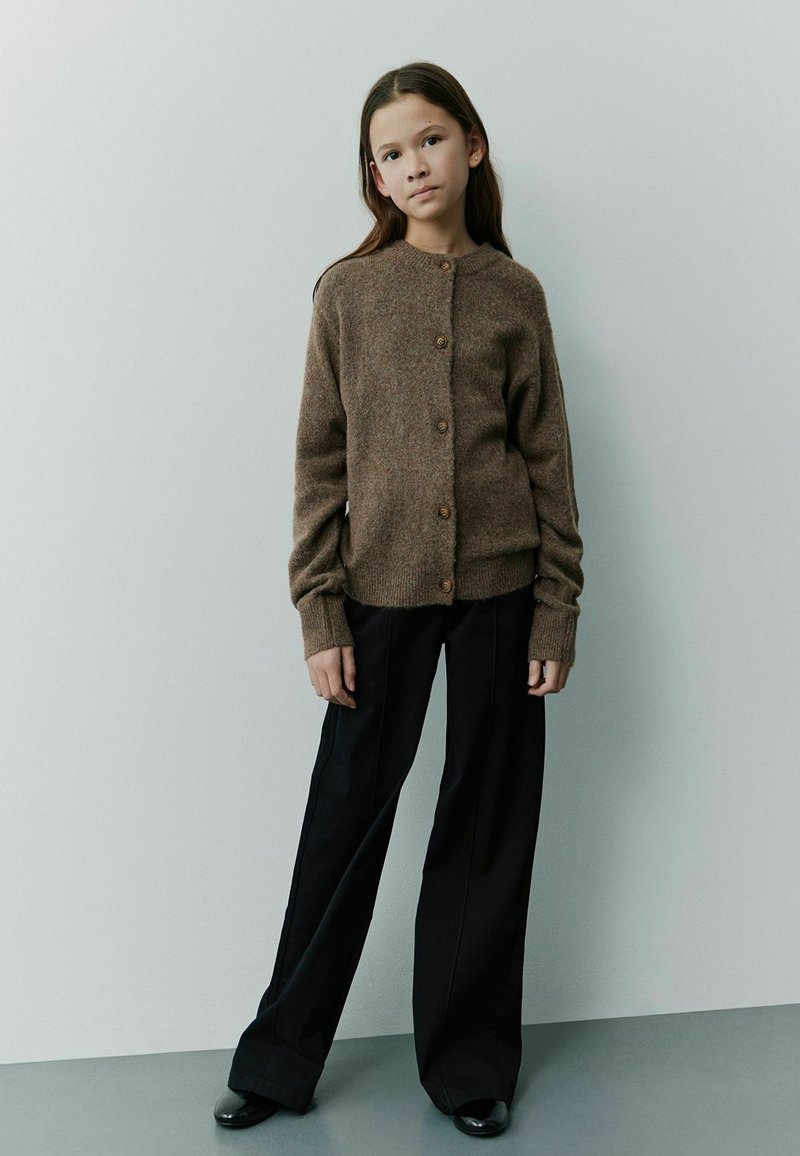 Braune, gestrickte Strickjacke mit Knöpfen, runder Ausschnitt, gerippte Bündchen. Kombiniert mit schwarzen, weit geschnittenen Hosen und schwarzen Schuhen.