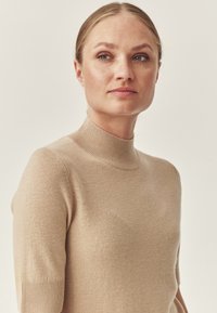 Maglione aderente beige con collo alto e maniche corte, realizzato in un tessuto morbido. Il tessuto appare liscio con una sottile texture.