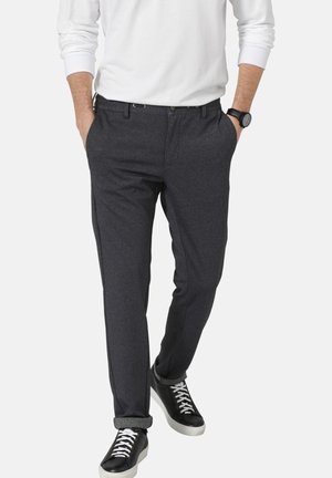 Kollargraue Hose aus strukturiertem Stoff, mit klassischem Schnitt und vorderen Taschen, kombiniert mit schwarzen Turnschuhen mit weißen Schnürsenkeln.