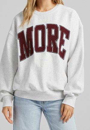 Kobieta ubrana w jasnoszarą bluzę z dużymi bordowymi literami układającymi się w słowo „MORE” na piersi oraz w jasnoniebieskie dżinsy.