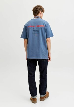 Moški v modri majici "Royal Denim", temnih zavihanih kavbojkah in rjavi obutvi, stoji z obračnim pogledom stran, na enobarvnem ozadju.