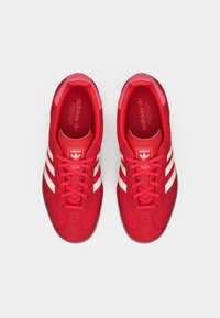 Κόκκινα αθλητικά παπούτσια Adidas με άνω μέρος από σουέτ και δίχτυ, με τρεις λευκές ρίγες, κλείσιμο με κορδόνια και υφή λεπτομέρειες στη γλώσσα.