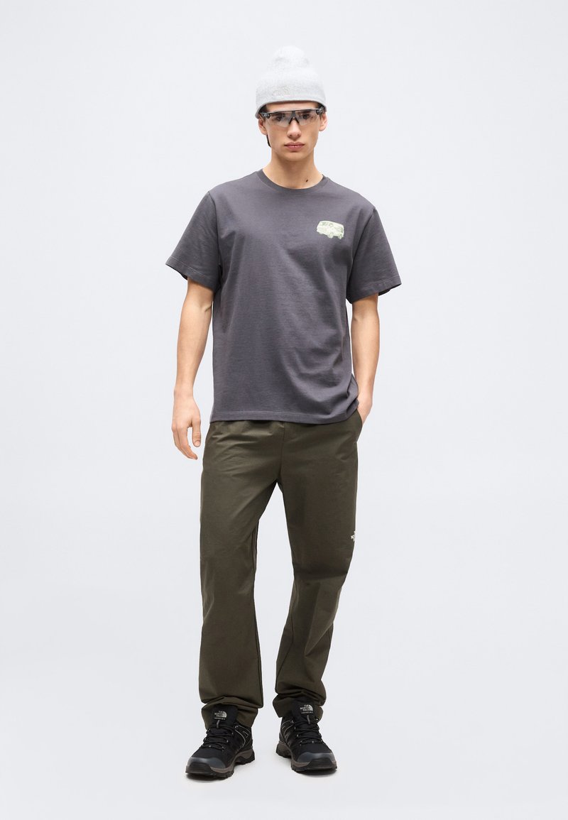 Junger Mann mit weißer Strickmütze, klaren Schutzbrillen, grauem T-Shirt mit kleinem Motiv, olivgrüner Hose und schwarzen Sneakers steht vor einem schlichten Hintergrund.