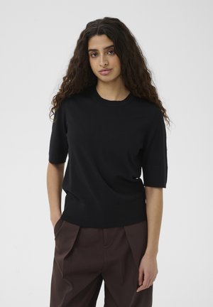 Woman with long curly hair wearing a black short-sleeve top and dark brown pants, standing with one hand in pocket against plain background.