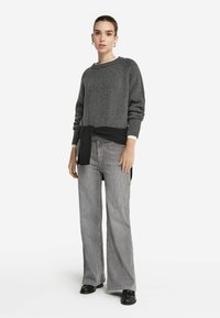 Grauer Strickpullover mit gerippten Ärmeln, über einem schwarzen Oberteil getragen. Kombiniert mit hellgrauen, weit geschnittenen Jeans und schwarzen Schuhen.