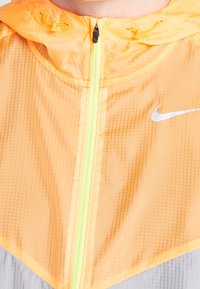 Rompeviento ligero en un brillante color naranja con un patrón texturizado, que cuenta con un zipper fluorecente verde y un logo de Nike en blanco.