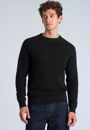 Homme portant un pull noir à côtes et un jean bleu foncé, debout face à l'avant, regardant légèrement sur le côté devant un fond clair et uni.