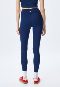 Mörkblå högmidjade leggings med en slät textur, med en liten logotyp på midjebandet. Kombineras med röda sneakers och vita strumpor.