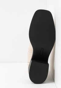 Chaussure en cuir beige au design épuré, surface texturée et semelle ronde noire. Présente des accents de lignes verticales sur la tige.