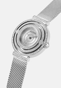 Zegarek na srebrnej siateczkowej bransolecie z okrągłą, ozdobioną kryształkami kopertą i okrągłą tarczą, z spiralnymi wzorami i logo marki na środku.