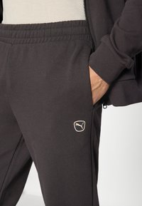 Pantalon de survêtement noir avec taille élastique, arborant un petit logo Puma doré sur le côté. Le tissu semble doux et lisse au toucher.