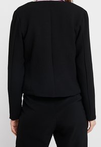 Femme vue de dos portant un blazer noir ajusté à manches longues et un pantalon noir assorti, sur un fond uni.