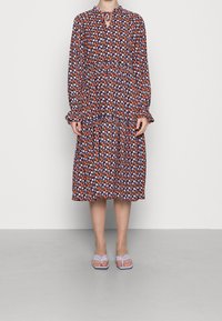 Vestido con estampado floral en negro, naranja y púrpura, con mangas largas, cintura fruncida y dobladillo con volantes. Combinado con sandalias de color púrpura claro.