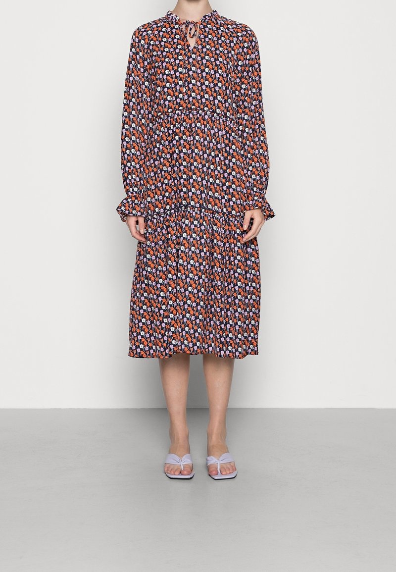 Vestido con estampado floral en negro, naranja y púrpura, con mangas largas, cintura fruncida y dobladillo con volantes. Combinado con sandalias de color púrpura claro.