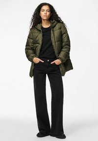 Olīvzaļš pūkains jaka ar kapuci, kas iezīmējas ar iešūtu dizainu un elastīgām piedurknēm, apvienota ar melniem platajiem džinsiem un melnām kurpēm.