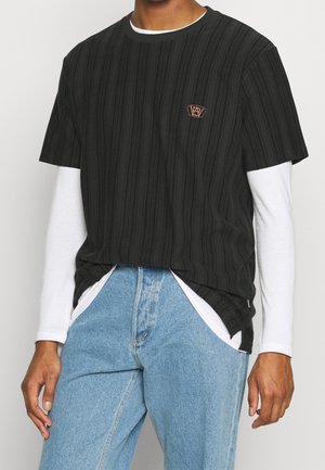 Personne portant des vêtements superposés : t-shirt noir à manches courtes sur chemise blanche à manches longues et jean en denim bleu clair, debout les mains détendues.