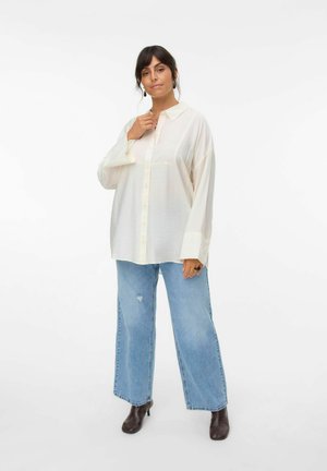 Camicia bianca oversize con colletto, maniche lunghe e chiusura con bottoni, abbinata a jeans a gamba larga chiari e sfilacciati e stivaletti alla caviglia marroni.