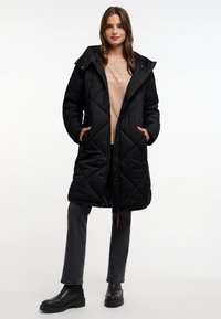 Zwarte gewatteerde pufferjas met een capuchon, voorzien van zijzakken en een ritssluiting aan de voorkant. Draag het over een beige top en grijze jeans, met zwarte laarzen.