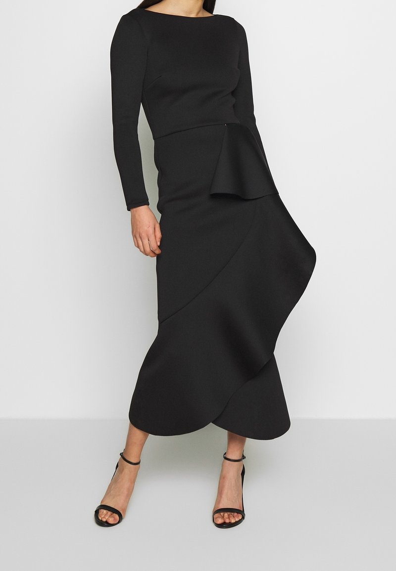 Femme portant une longue robe noire à manches longues avec une jupe asymétrique à volants, associée à des talons noirs avec bride à la cheville.