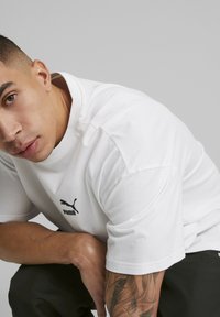 Hvid kortærmet T-shirt lavet af bomuld med rund halsudskæring og et sort Puma-logo på venstre bryst. Glat tekstur, afslappet pasform.