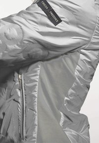 Zilveren donsjas met een zak met rits, textuurpatronen en een logolabel. Het materiaal is glanzend en glad, met een gevoerde binnenkant.