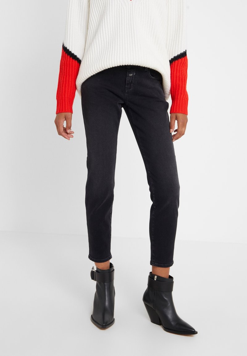 Jean en denim noir à coupe slim, revers roulés, associé à des bottines noires. Porté avec un pull en laine blanc et rouge.