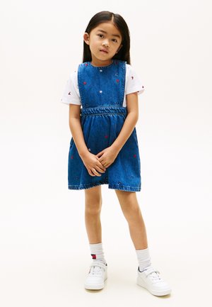 Mladé dievča stojí, oblečené v modrých rifľových šatách s malou červenou a bielou kvetinovou výšivkou cez biele tričko a biele tenisky.