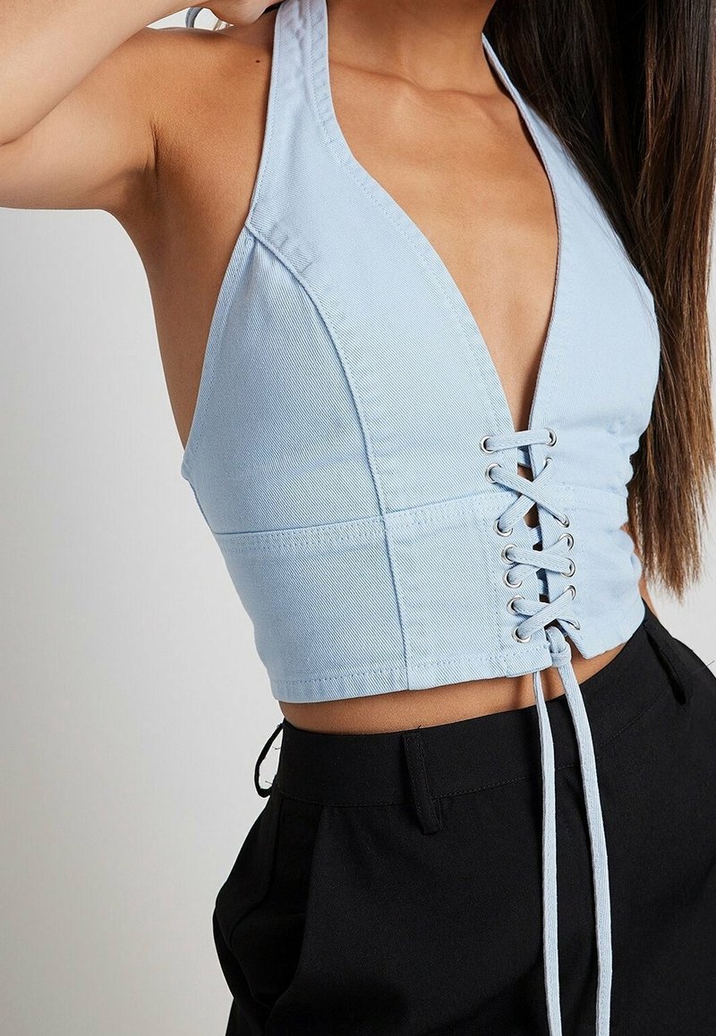 Femme portant un haut dos-nu bleu clair avec un détail lacé à l'avant et un pantalon noir taille haute, coiffure longue et droite.