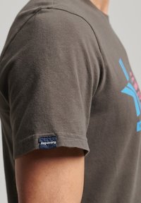 Harmaa puuvillapaita, jossa on pyöreä kaula-aukko, lyhyet hihat ja sininen "Superdry" -etiketti hihassa. Edessä on näyttävä värikäs graafinen kuvio.